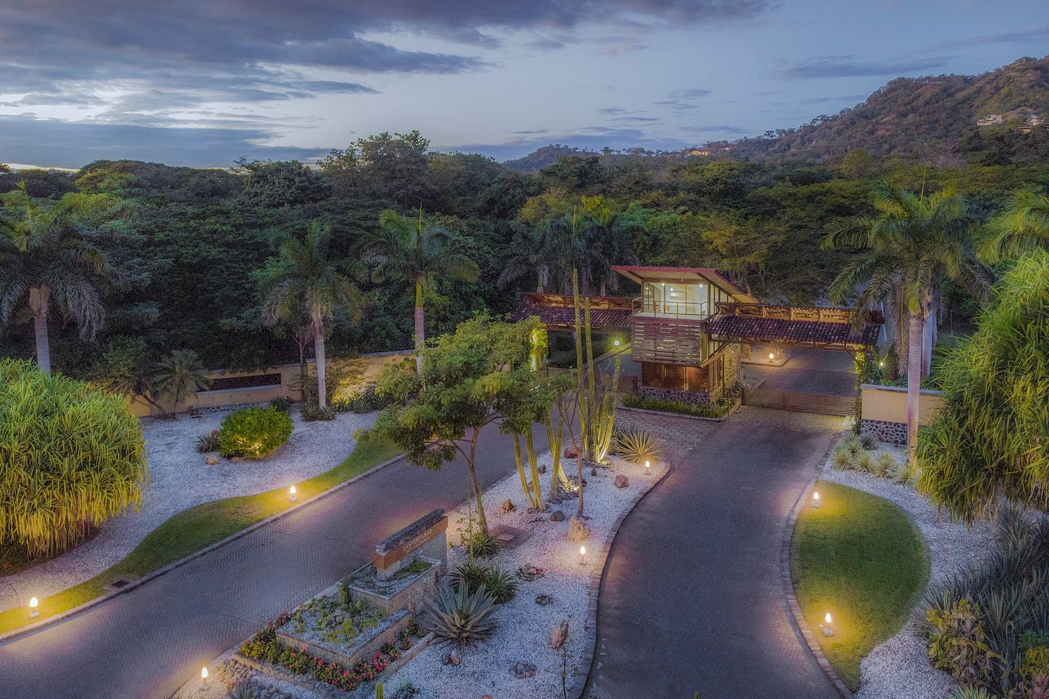 A lit community entrance gatehouse at dusk, uplit palms flanking the drive