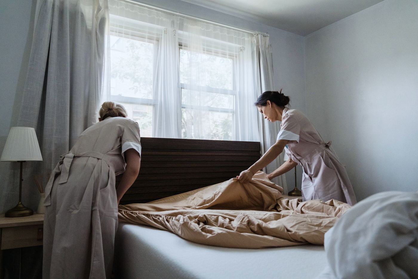 Two housekeepers in matching uniforms making a bed by a bright window
